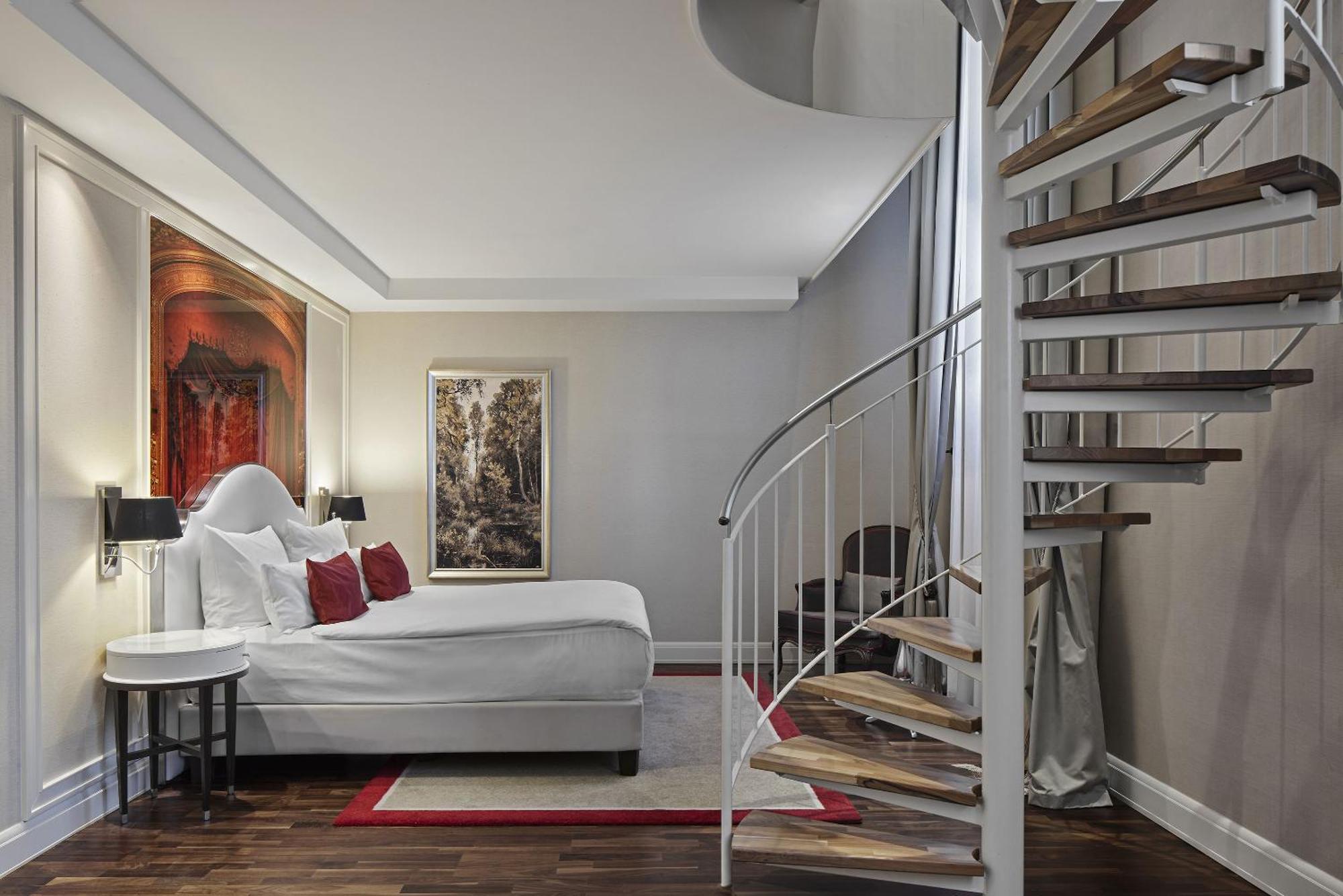 Titanic Gendarmenmarkt Berlin Extérieur photo The photo showcases a modern bedroom featuring a queen-sized bed with a white comforter and decorative red pillows. The room has a stylish and contemporary design, with hardwood flooring and soft lighting. On the walls, there are two pieces of art: o